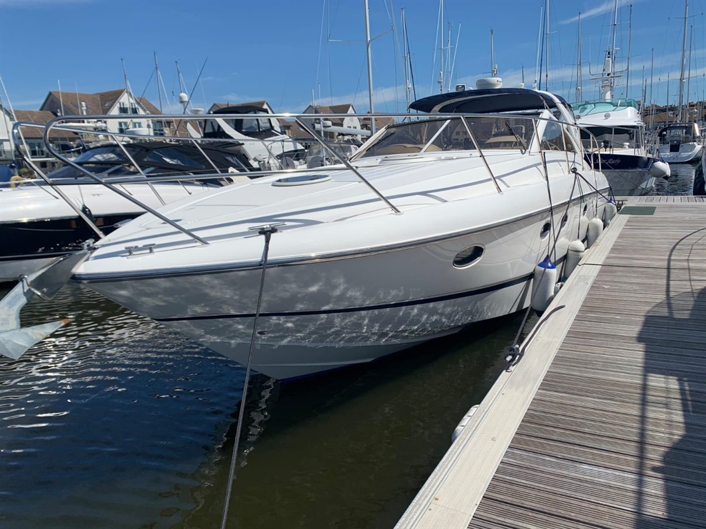 A white boat at a marina, docked beside other vessels, with a clear blue sky and sunlight reflecting on the water.